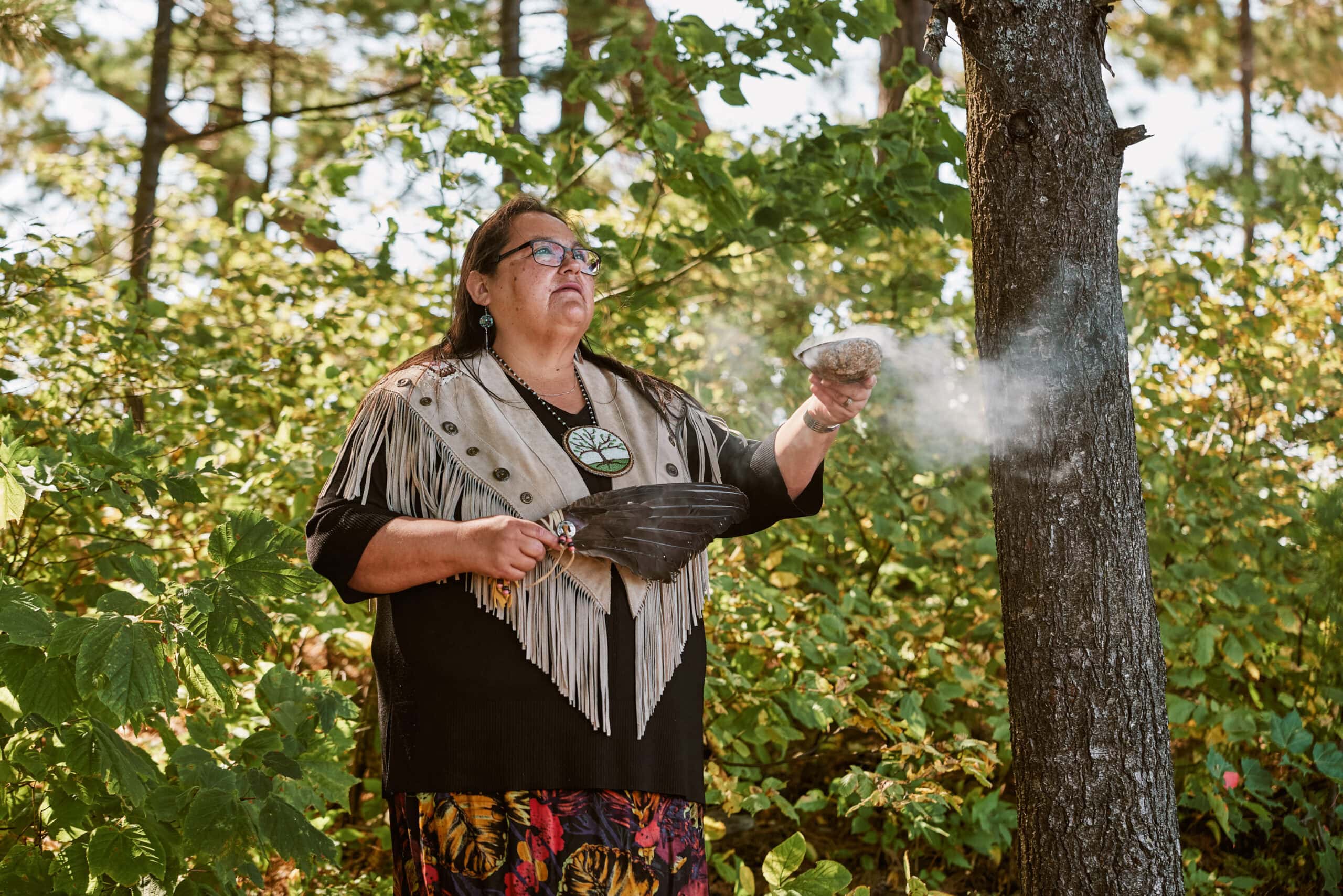 An Anicinabe woman in ceremonial attire stands in front of trees with an eagle feather performaing a smudge ceremony with a bowl made of a seashell.