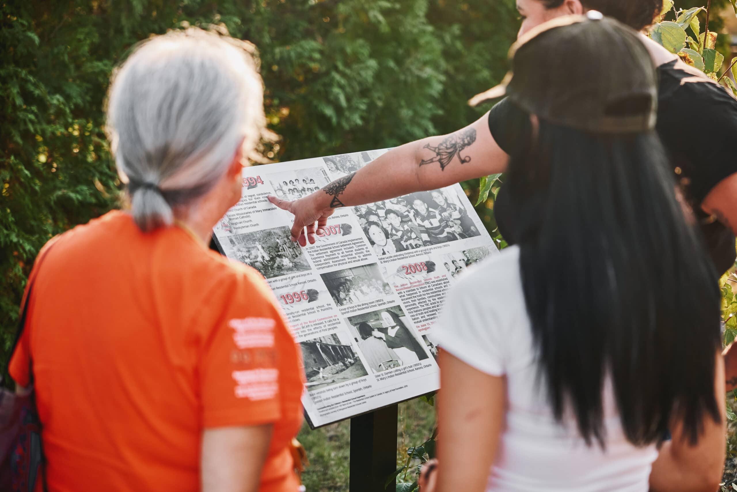 A woman points to an image on an outside exhibit about Residential Schools while an Elder and another young woman look on.