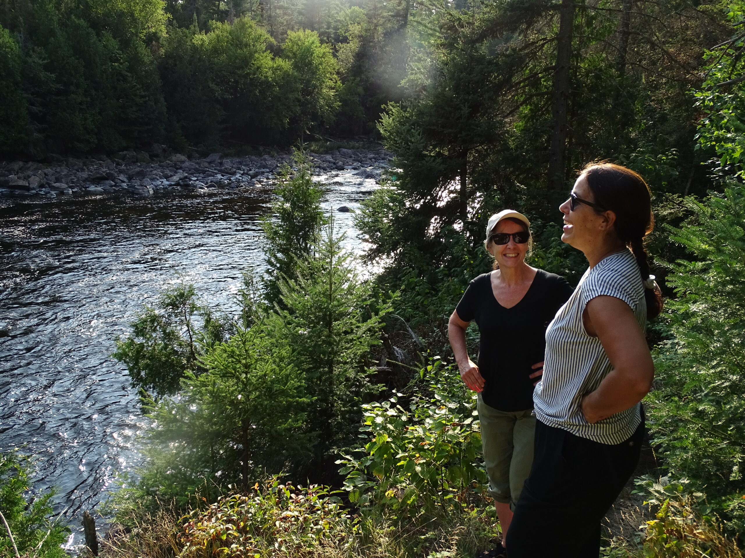 Two women laugh and smile in Park d'Opemican near a river on a sunny day.