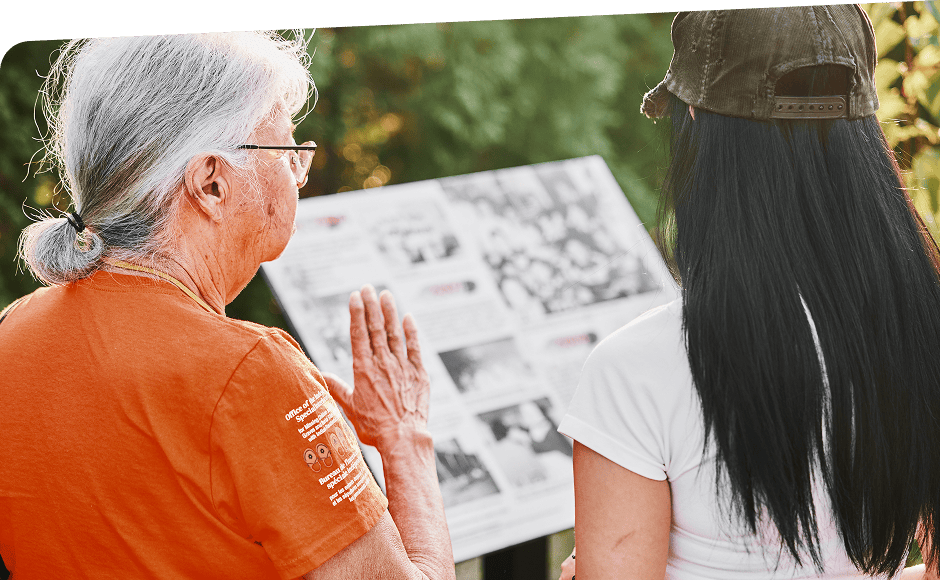 Subtract An Anicinabe Elder wearing an orange shirt speaks to a young woman in front of an outdoor exhibit about Residential Schools.